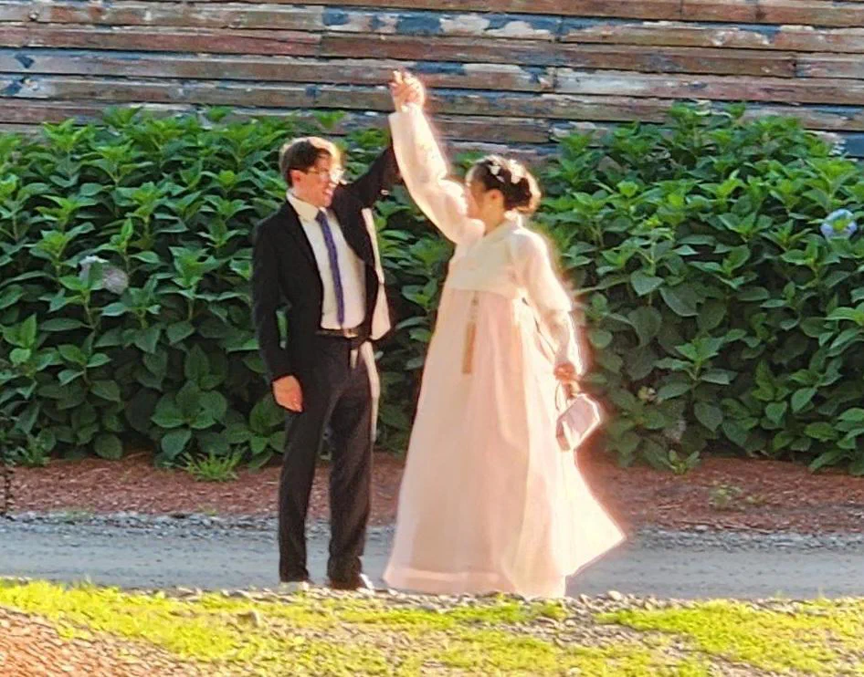 My husband and me mid-dance outdoors, hands raised together, in front of a hedge of hydrangeas.
